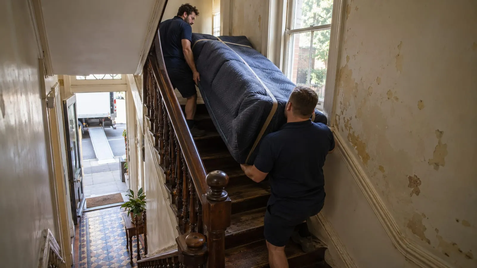 Two removalists carrying a wrapped sofa up the narrow turning staircase of a Sydney terrace house